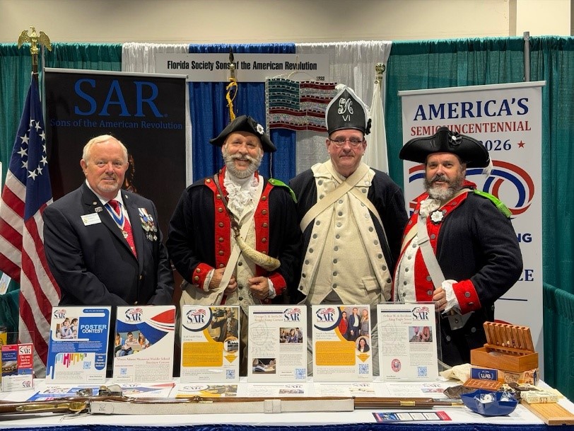 Four Florida Society SAR members behind an exhibition table spread with youth contest flyers. One wears a blazer with SAR medals; three wear Continental Army uniforms and tricorn hats. Banners behind them read Florida Society Sons of the American Revolution and America's Semiquincentennial 2026.